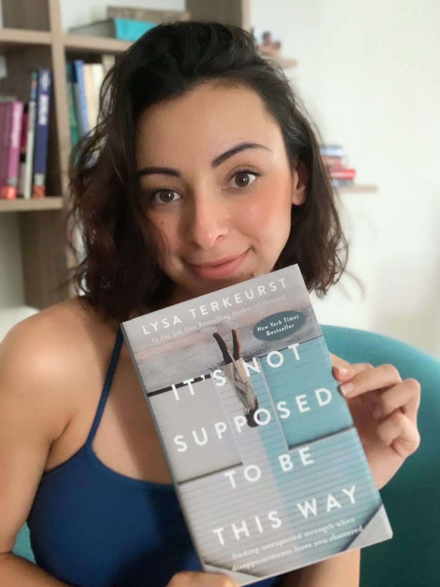 Arja, a woman with short, curly brown hair, poses with a book by Lysa Terkeurst titled, "It's Not Supposed to Be This Way" She wears an blue camisole and poses in front of a bookshelf.
