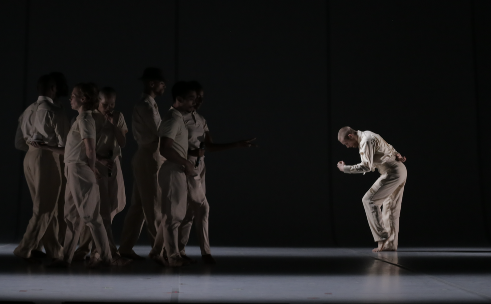 One dancer, well-lit on a stage, leans over apart from a group in shadow