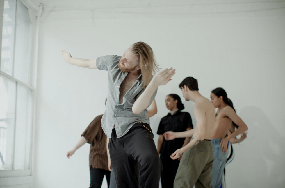 With long blond hair, Tedholm cuts his elbow to his waist, bending toward it with one knee lifted. Dancers behind him in the white space congregate