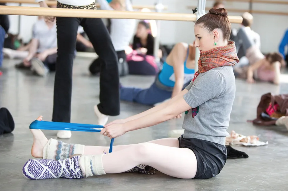 A ballet dancer sits below the barre, surrounded by other dancers in the background warming up. She has a bare foot in an exercise band, flexing her foot into it.