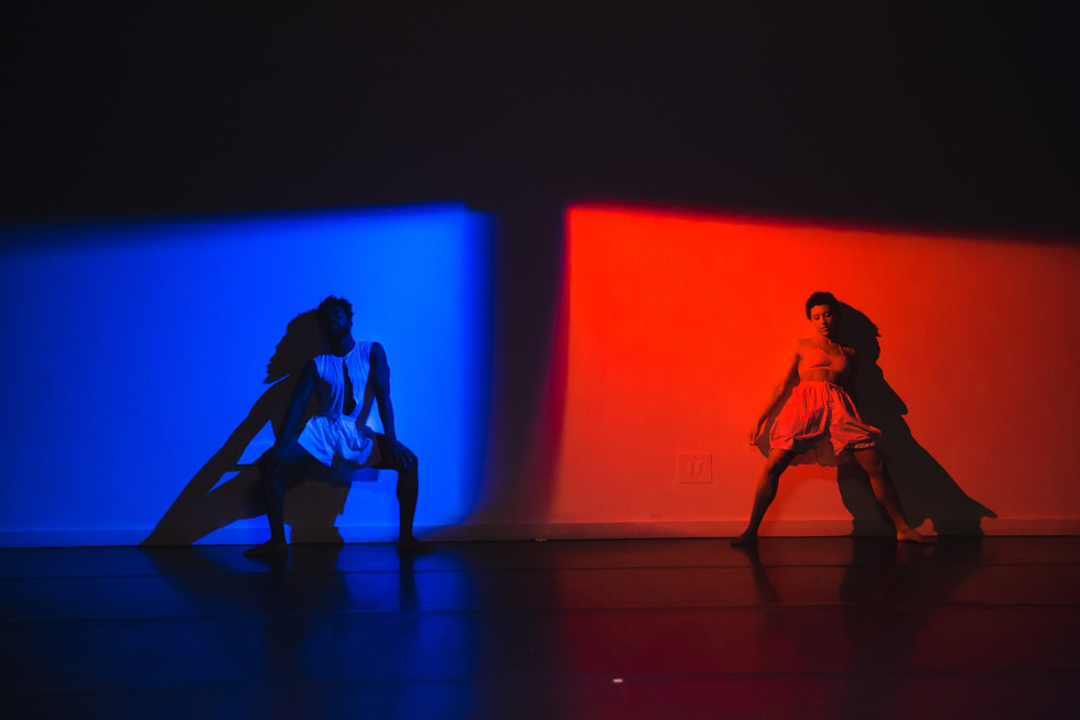 A dark-skinned man and woman lean against an upstage wall with their upper bodies as they pliu00e9 in second position. The man and the wall behind him are bathed in blue light; the woman and her half of the space in red light.