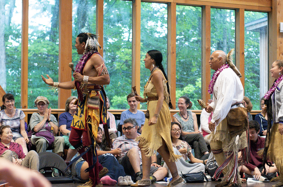 A procession of performers in Native American clothing walks across the studio, holding instruments and singing, as onlookers watch from the sides.