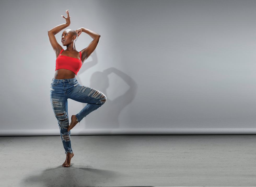 Gabrielle Hamilton, a dark-skinned young woman with a shaved head, dressed in high-waisted, ripped jeans and a red crop top, balances on relevu00e9 in retiru00e9. She looks to the left, arms angularly framing the space around her head.