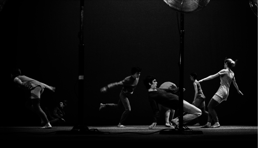 In a black and white image, seven dancers are scattered, for the most part mid-run. One hinges almost to the ground, their fingertips just brushing the floor. Another is caught either mid fall or mid rise, tipping off balance backwards to the floor.