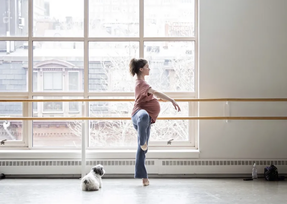 A pregnant dancer stands in passe at the barre, a small dog behind her.
