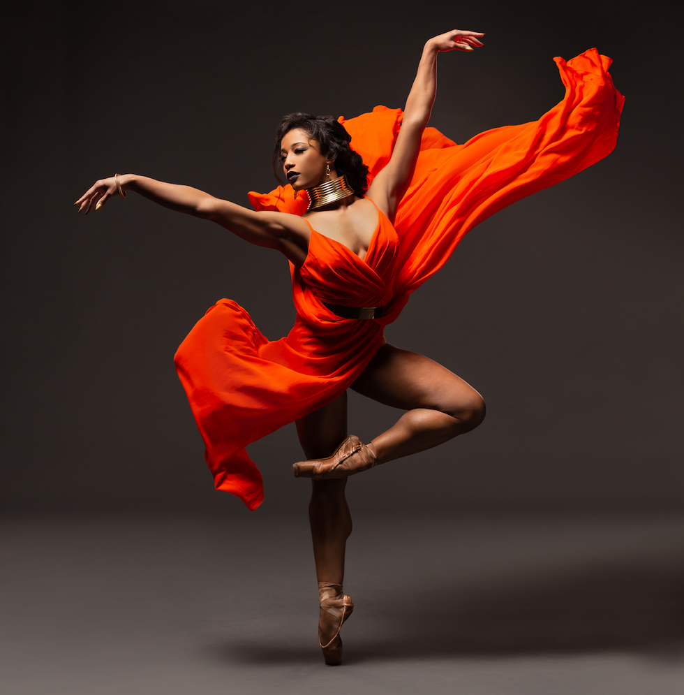 Parina stands on pointe in passe front, and orange dress flowing around her on a grey backdrop.