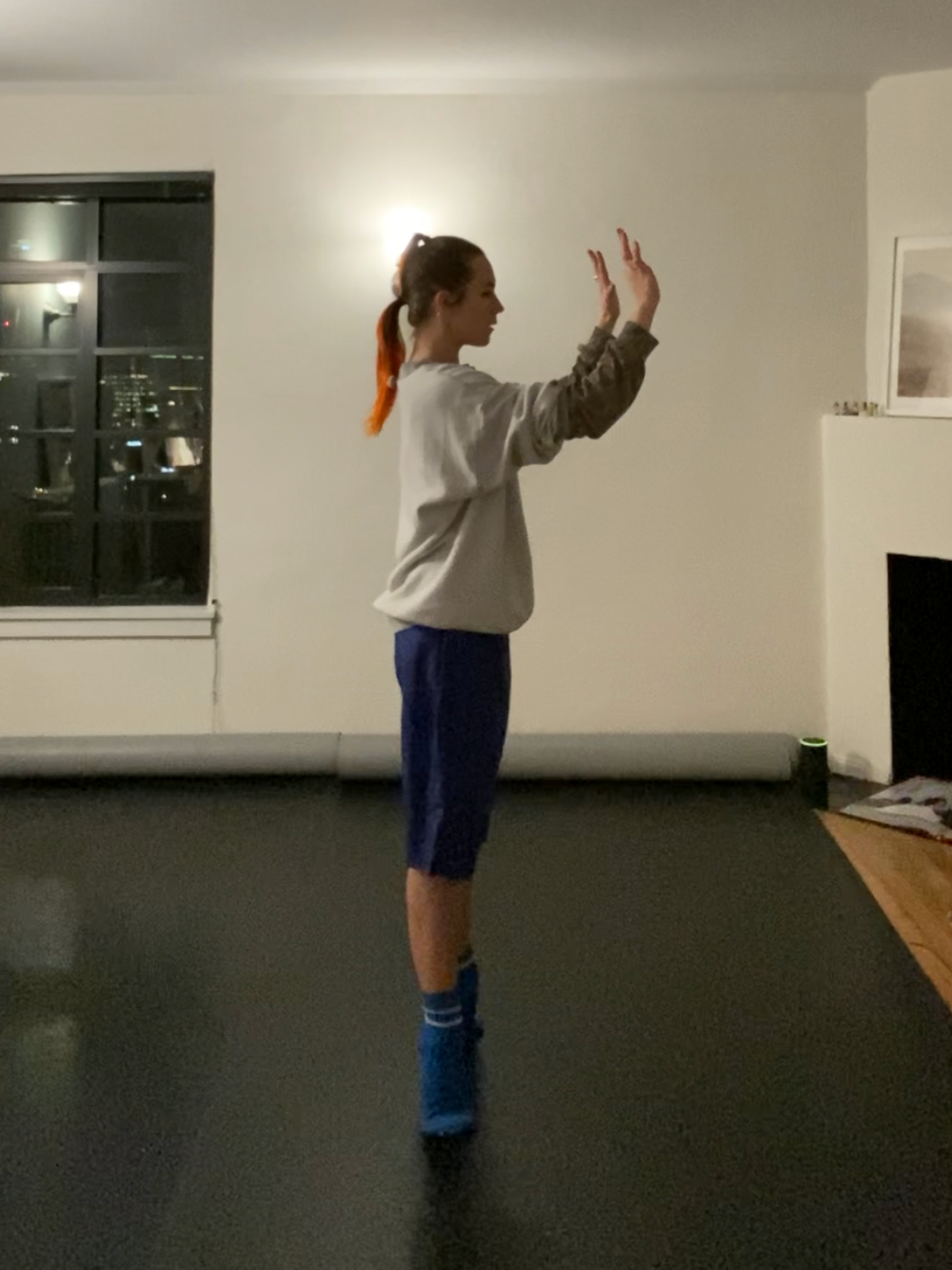 Wallich, a woman with her hair in a low ponytail, stands in her apartment. Two gray marley rolls are laid out beneath her, and there is a white wall behind her. She wears blue socks, dark blue shorts and a grey sweatshirt. She balances in relevu00e9 with her arms placed in front of her, palms facing outward.