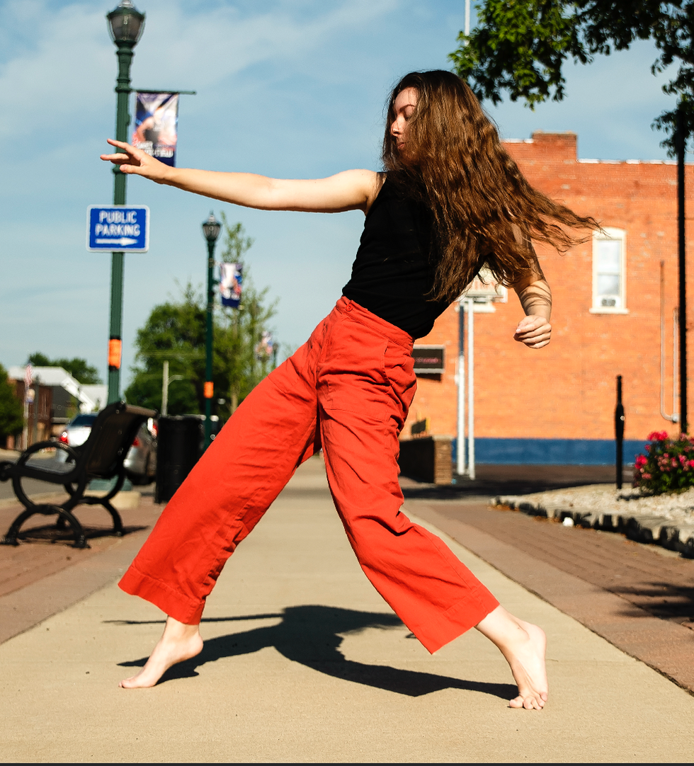 Emily Pierce, barefoot and waring red trousers and a black top, closes her eyes as she moves on an outdoor walkway. She hinges back, away from her outflung right arm, long brown hair swinging behind her.