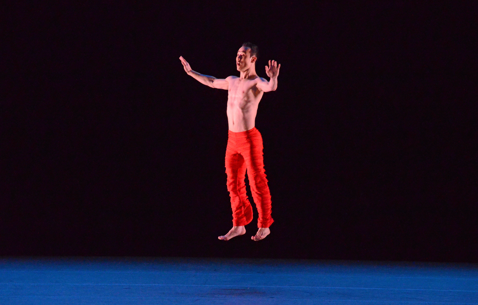 A bare-chested Asian man jumps straight up into the air on a dark stage, his expression surprised, arms gently bent out to the sides