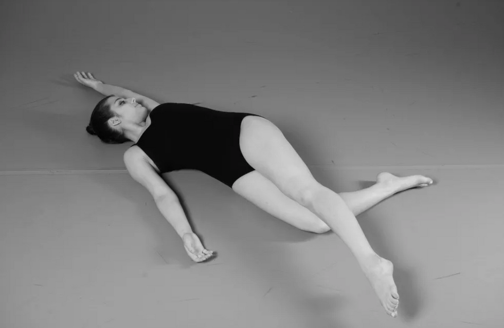 A dancer lies on the floor with one arm above her head and the leg on the same side reaching diagonally across her body.