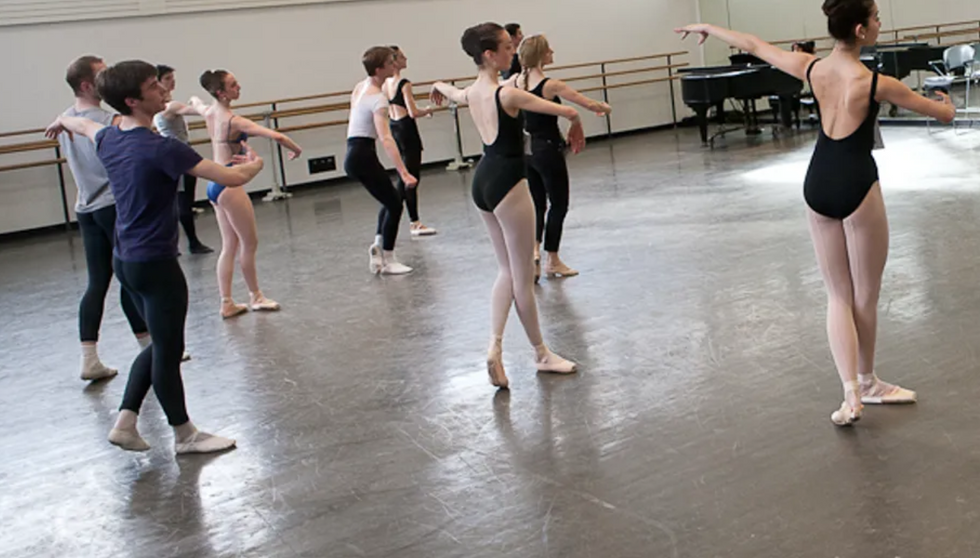 A group of ballet dancers who seem to be learning choreography in a dance studio. They are in rehearsal wear and look like they are marking movement being taught at the front of the room.