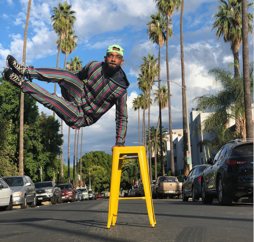 Jamal Josef jumps sideways on a palm-tree lined street, balancing his weight on a yellow stool