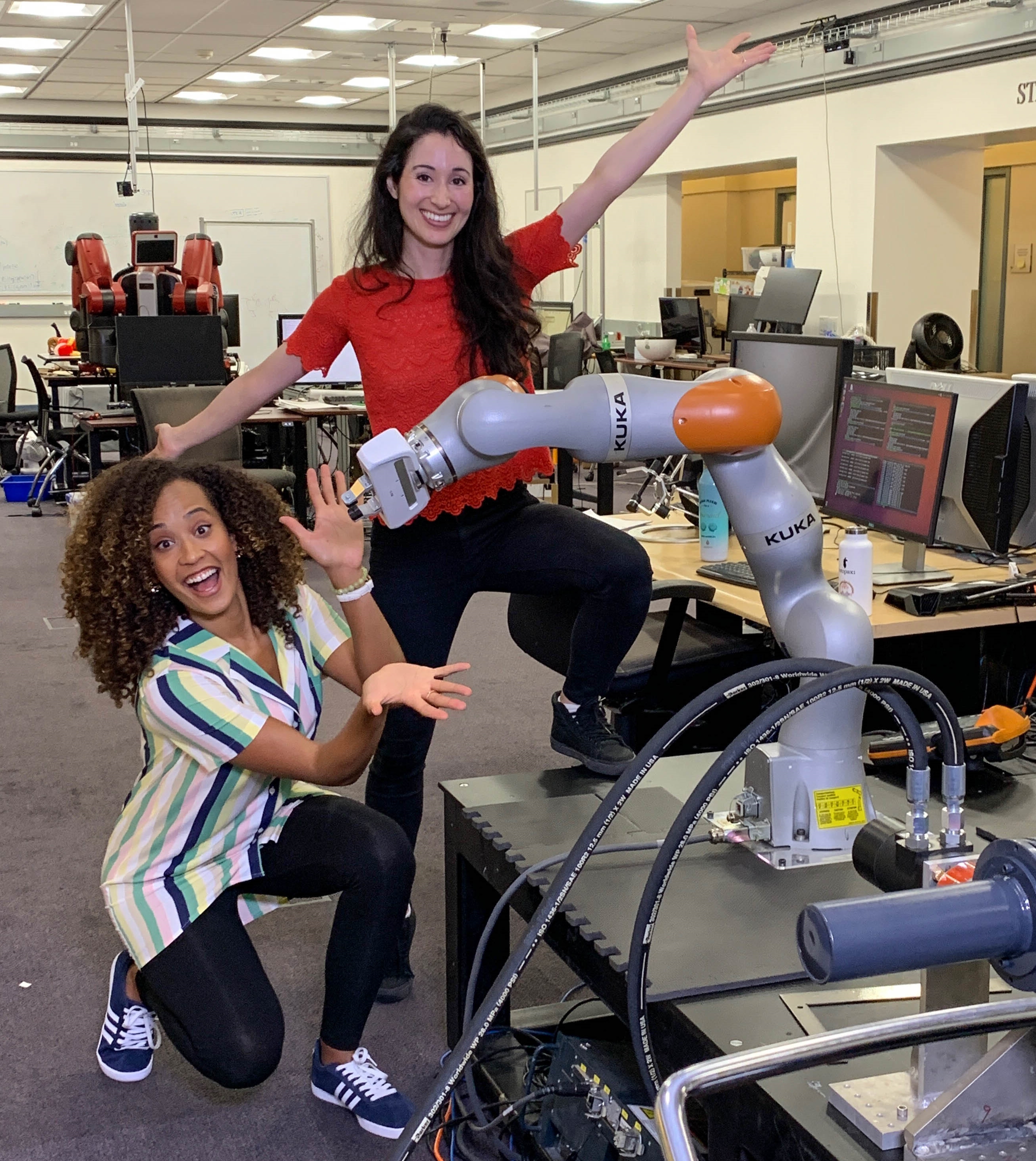 Danni Washington, a Black woman with curly hair, kneels next to a robotic arm, while Catie Cuan, stands next to her. Both women are pointing toward the robot with enthusiastic gestures.