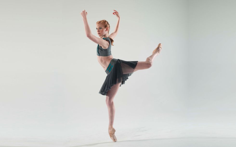 A red-headed ballerina balances in a forced arch back attitude en pointe. Her arms are bent at the elbows and in slight opposition to the twist of her open attitude so her forearms frame her face.