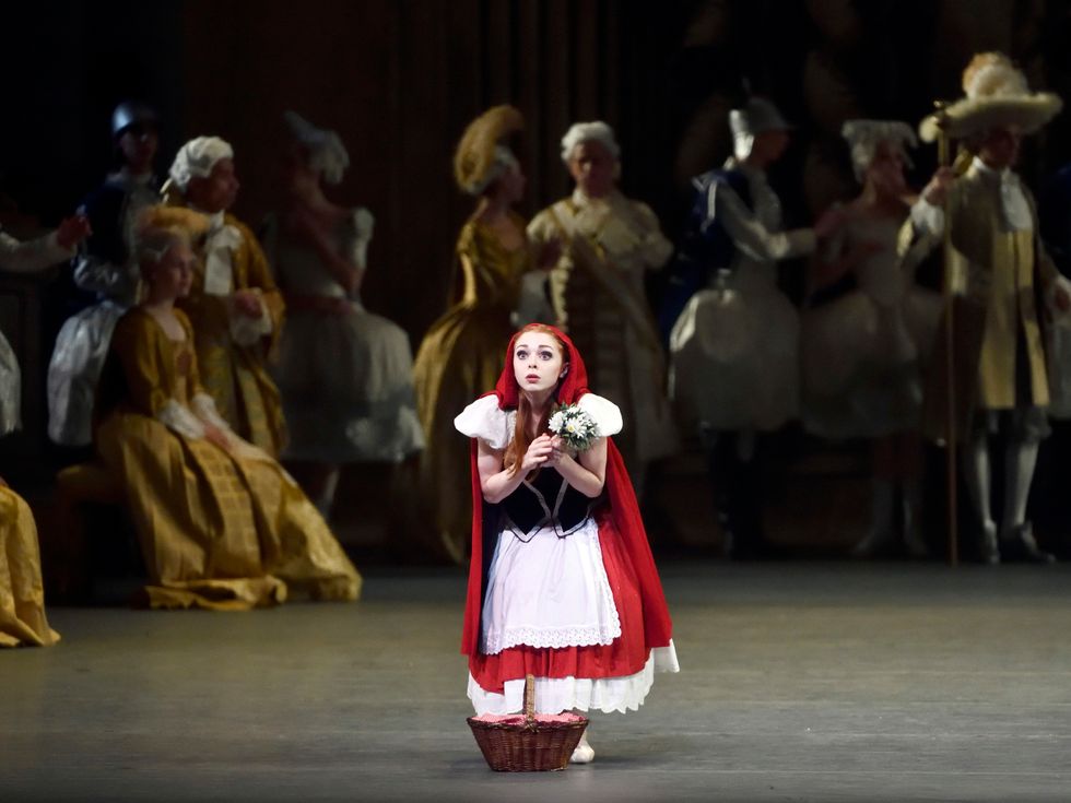 A short, red-headed ballet dancer dressed as Little Red Riding Hood stares out at the audience with wide eyes. She clutches flowers in both hands as she peeks out from beneath her red robe, a wicker basket by her feet.