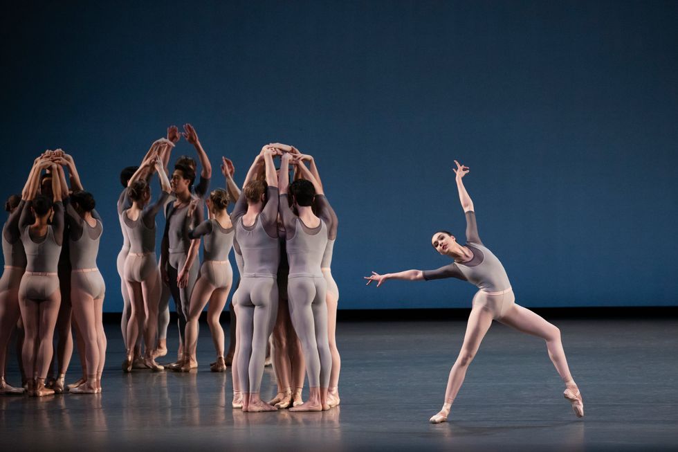 To the left, small groups of dancers dressed in gray leotards and tights (pink for the women, gray for the men) cluster in small circles facing in, hiding a dancer in the center from sight as they clasp their hands overhead. To the right, a long limbed dancer with striking features leans with one pointe-shoe clad foot hovering just above the stage in what is almost an open lunge. She arches toward her back leg as her arms form a slightly over-extended 'V' above her shoulders.