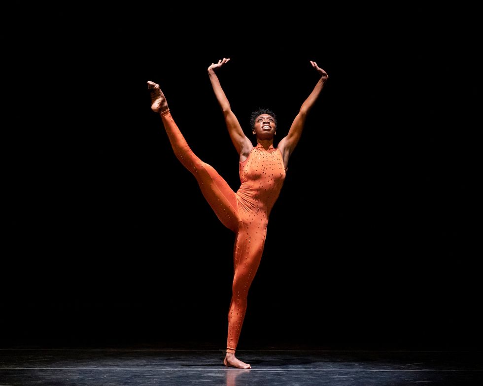 A dark-skinned woman wearing an orange unitard smiles as she extends her right leg high to her side, her supporting leg turned out and steady. Her arms stretch into a "V" overhead that matches the angle of her extended leg, wrists flexed and palms upturned. Her chin and eyes are lifted.