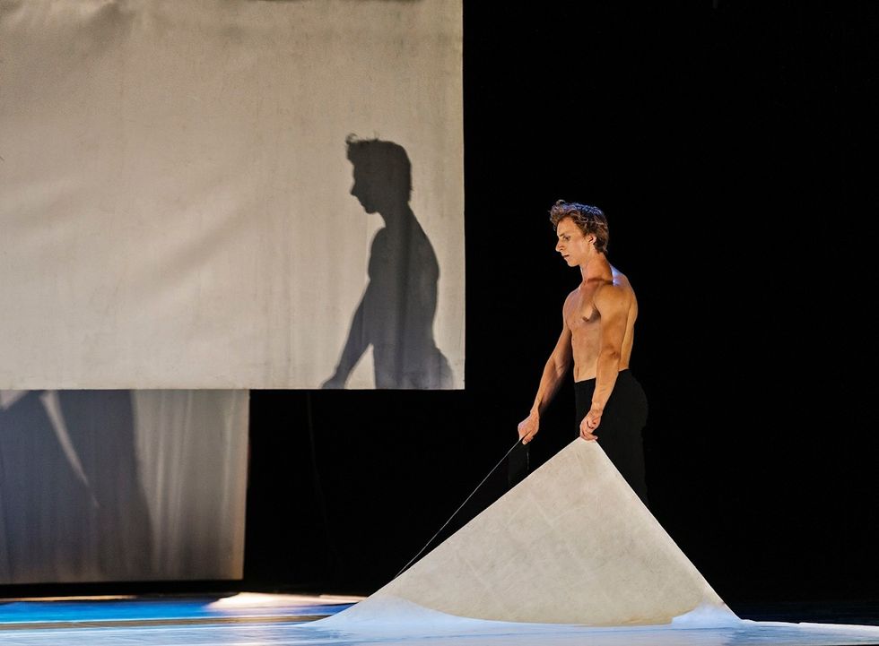 A bare-chested male dancer lifts the corners of two panels of marley, arms raised slightly in front of his sides. His shadow is cast on a piece of fabric just upstage.