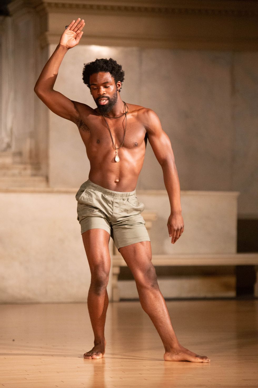 A long-limbed, bare-chested black man stands with one hand raised. He sits into his right hip, left leg extended slightly in front of him but with the foot flat on the floor. His gaze is downcast as his right arm, bent at the elbow, rises alongside his head.