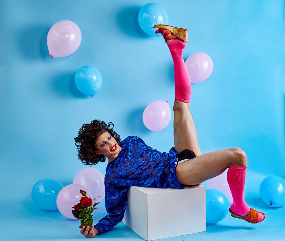 Blue and pink balloons that read "It's a boy!" or "It's a girl!" are scattered around a blue space. In the center, Yergens reclines on a white block. He is wearing a wig of wild brown curls and dramatic makeup, a blue flower patterned shirt, underwear, hot pink knee-high socks, and wedge heeled sandals. His left leg kicks straight in the air, as though he just sent the balloons flying, while his right hand rests on the floor as it holds roses.