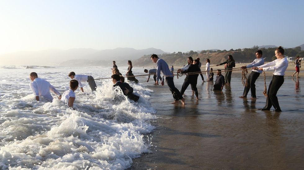 More than a dozen performers stand on a sandy beach, waves crashing in, hills in the background fading into mist. Some of the performers hold long ropes, which are wrapped around the waists of others who are facing or partially submerged in the oncoming surf. Others walk in of their own volition, while still others watch from their knees.