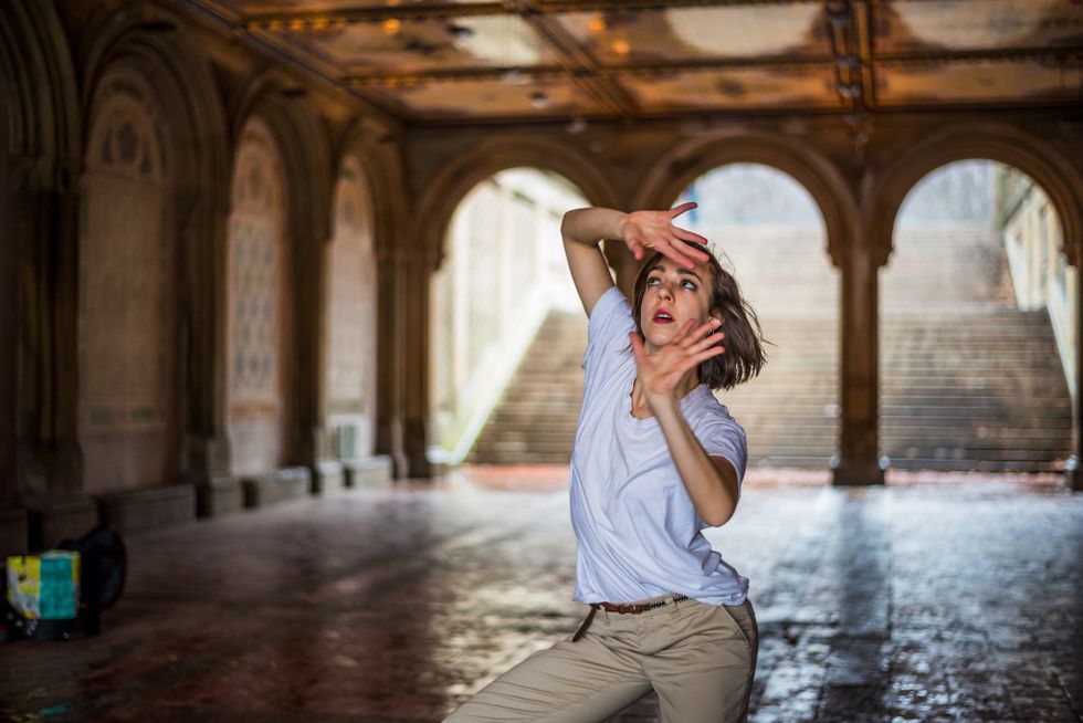 A woman in a loose white t-shirt and khaki pants stands underneath an opulent archway in Central Park. She sits into her left hip, right knee cocked, as her open palms frame her face from above and below. Her eyes glance past her upper hand.