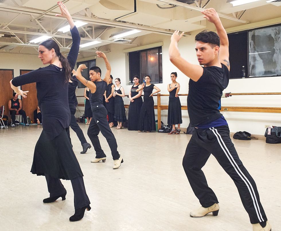 Dancers dressed in all black rehearse in a studio. Beltran, right, imitates the pose of the woman demonstrating choreography, his left leg stretched to one side as he pliu00e9s on his right, upper body twisting slightly away from the extended leg as his arms rise to curve around his head. He stares intently forward.