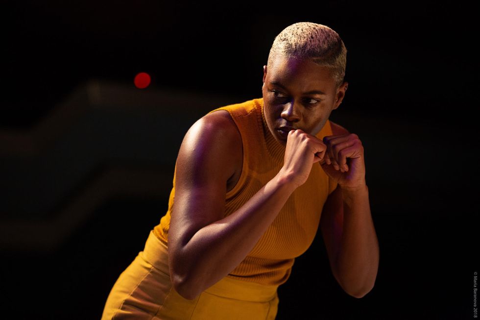 A dark-skinned woman with cropped blonde hair stoops over slightly, fists raised in front of her mouth. She looks warily toward the left.