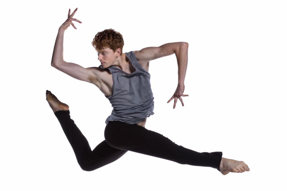 A red-headed male dancer leaps. His right leg crosses straight in front, while his left bends into a parallel attitude behind him. He is twisted toward his back leg, elbows bent at 90-degree angles with his back forearm stretching up and front forearm pointed down.