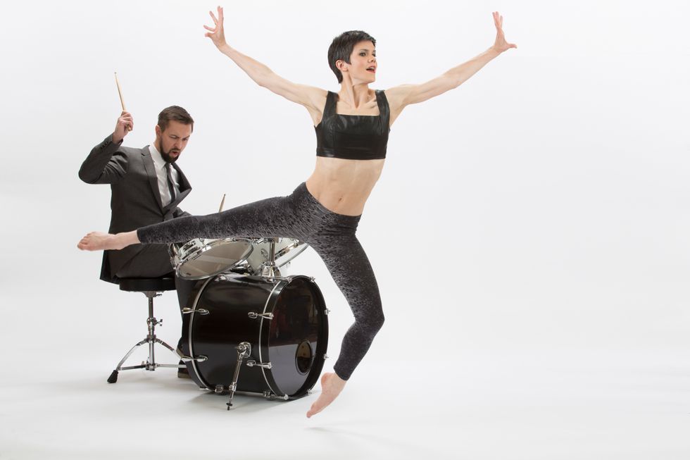 A besuited man is seated at a drum kit in a white space, stick raised to strike in his right hand. In the foreground, a woman with a tightly cropped pixie cut, wearing textured leggings and a crop top, leaps. Her left knee is bent, foot pointed just above the floor, as her right leg stretches behind her. Her arms are flung open as she looks energetically toward the right.