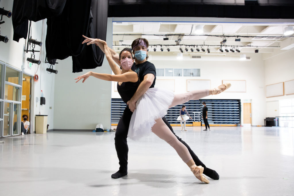 In masks and ballet rehearsal wear, two dancers strike a pose recognizable from the Black Swan Pas de Deux. The ballerina hits a 90 degree third arabesque, shifted forward off of her center by the male dancer supporting her at the waist.