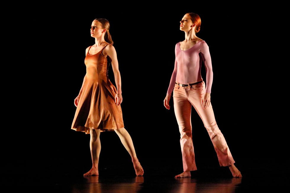 Two women, one in a dress and one in pants and a leotard, look off to the side of a dark stage