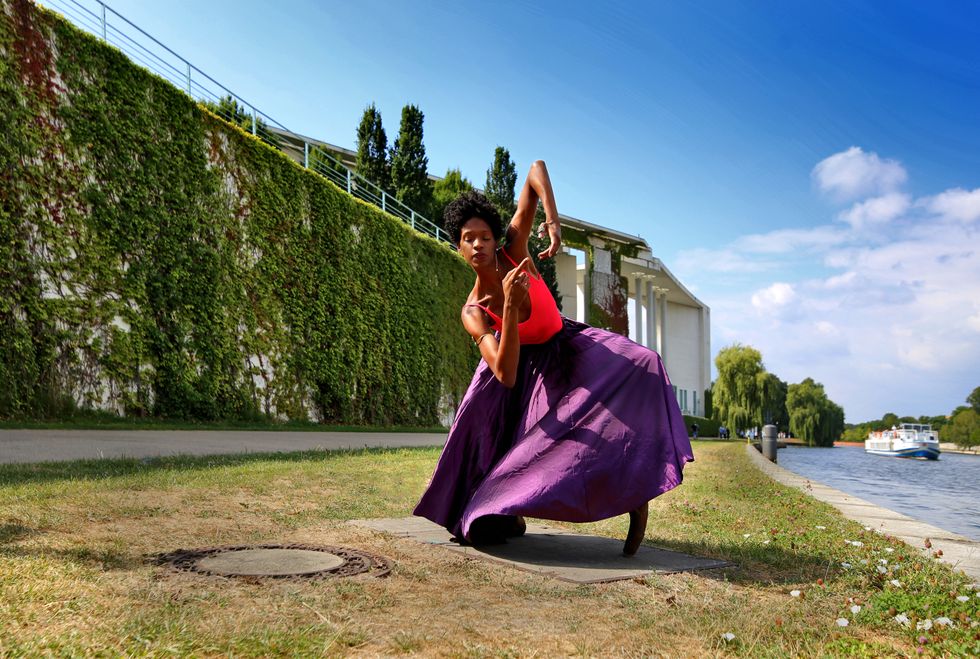 A black dancer squats in a purple skirt along the banks of a river