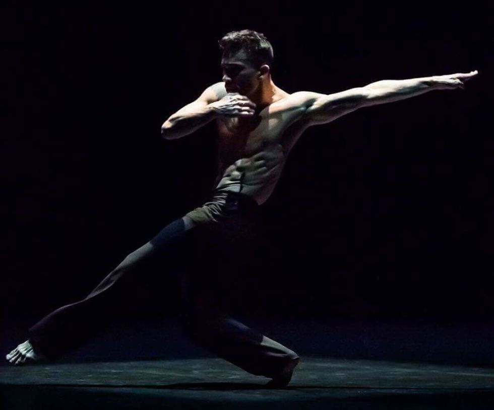 Jon Bond, barechested on a dark stage, reaches one leg far across his body while his arm reaches in the opposite direction