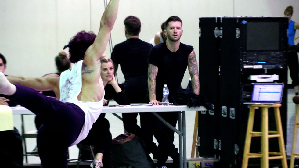 Travis Wall, a white man dressed in plain black clothing that reveals tattoo sleeves, stands behind a white table at the front of a mirrored studio. In the foreground, a male dancer with his back to the camera moves through arabesque.