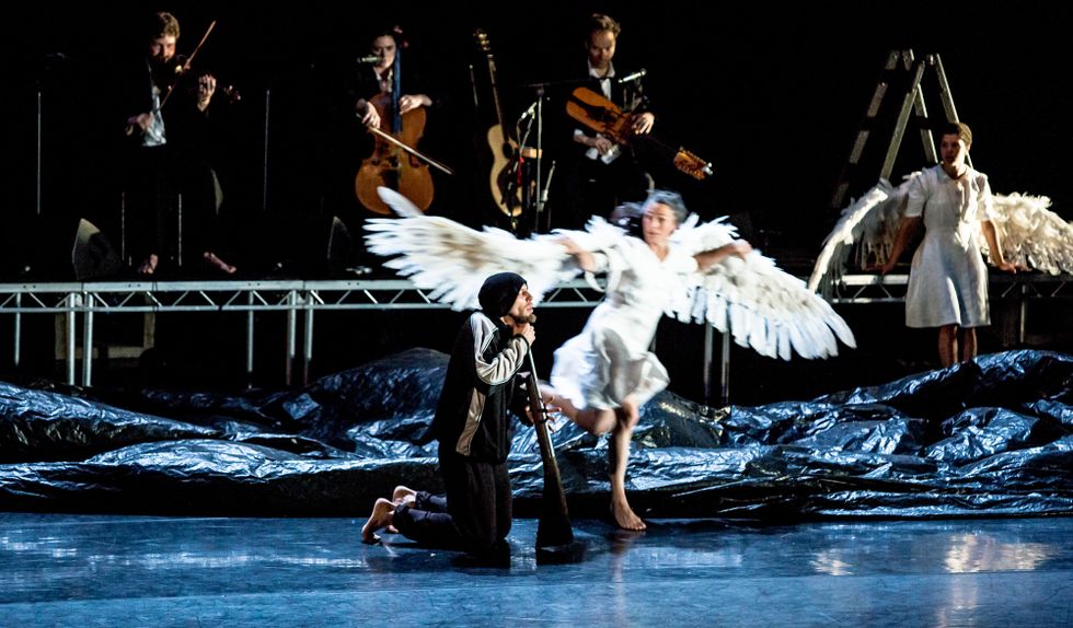 A man wearing a windbreaker and a beanie kneels, his chin pressed against the barrel of a shotgun. A blurry woman dressed in white with large wings runs past, looking back at him. Behind her, the stage is covering in a black, trash bagu2013like material. On a raised platform upstage, three barefoot, besuited musicians hold string instruments. Another woman in white with wings leans back against the musician's platform.