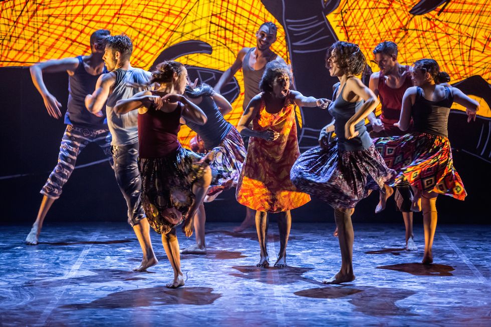 Eight barefoot dancers, half men and half women, dance barefoot around an older woman in an orange and yellow dress who seems to be mid-song or chant. The dancers wear colorful trousers and skirts and plain tank tops; they all look delighted.