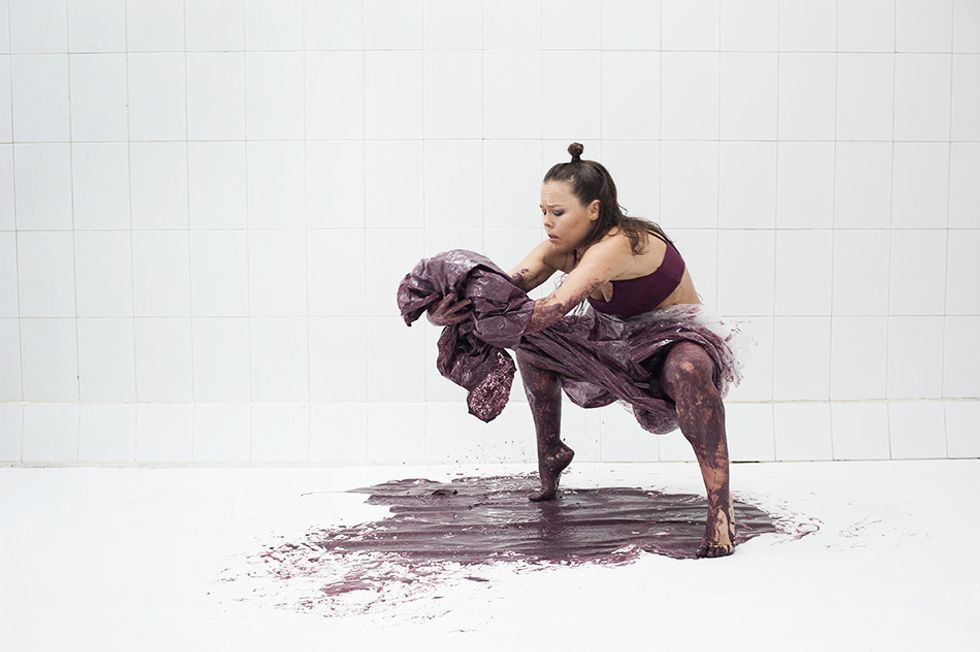A woman drips a puddle of plum-colored paint onto the floor of a clinical white room. Her arms cradle the bundle of her long, drenched skirt as she hunches over a deep second position pliu00e9.