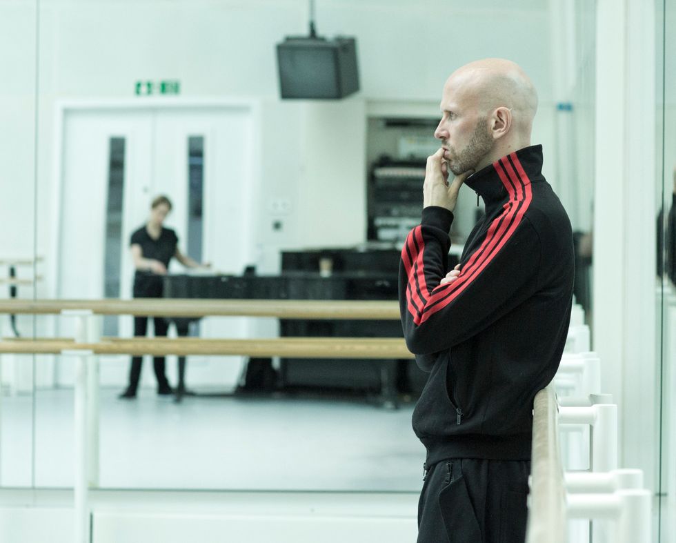 A white man with a shaved head and wearing a black tracksuit leans against a barre in profile, one hand raised to his mouth as he stares intently into the studio. In the mirror in the background, a woman looking at papers atop a piano.