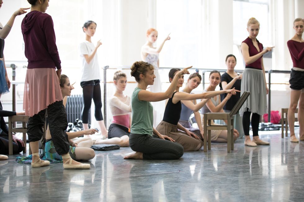 In a busy studio, a woman kneels, sitting back on her heels, with one hand resting in her lap and the other arm raised to 90 degrees and bent at the elbow, one finger pointing. Behind her, female dancers in pointe shoes and ballet slippers watch and imitate the gesture, some kneeling like Marston, others standing.