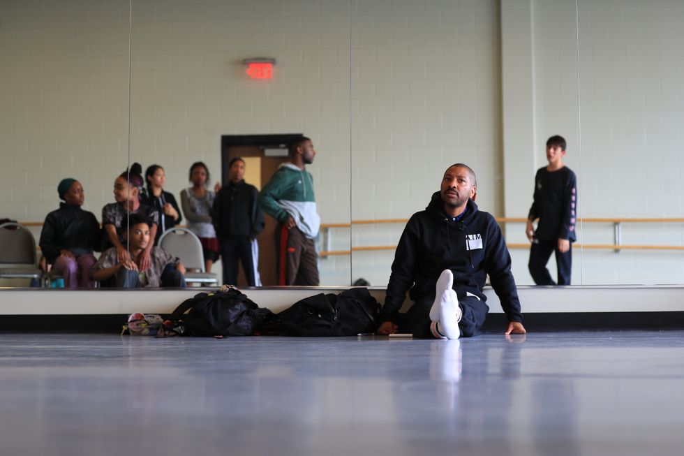 A dark-skinned man sits at the front of the studio, legs stretched out in front of him, back against the mirror. Out of focus in the mirror are dancers in a pause in rehearsal, all dressed in warm-up gear.