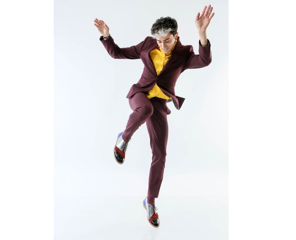 Teicher at a photoshoot in front of a light gray background, wearing a maroon suit, bright yellow shirt and silver, red and black tap shoes. He seems to be in the middle of a tap step, one leg raised in the air with the knee bent, arms casually flung in the air, looking down at his feet.