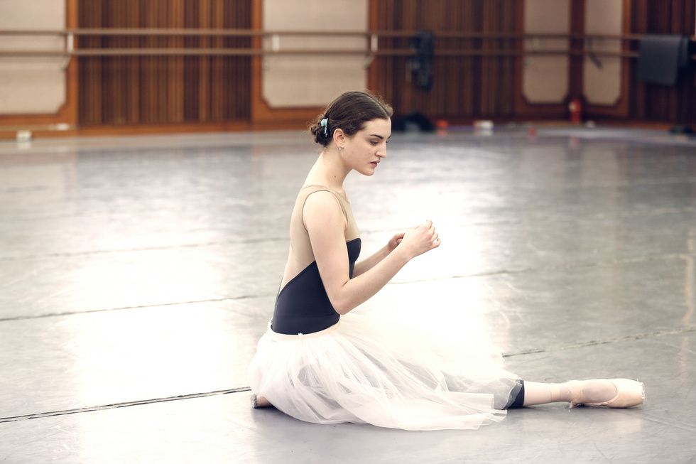 Womack in rehearsal in a studio, wearing a tan and blue leotard and a white romantic practice tutu. She kneels on the ground, her arms gesturing in front of her, eyes turned downward, as if rehearsing a dramatic moment.