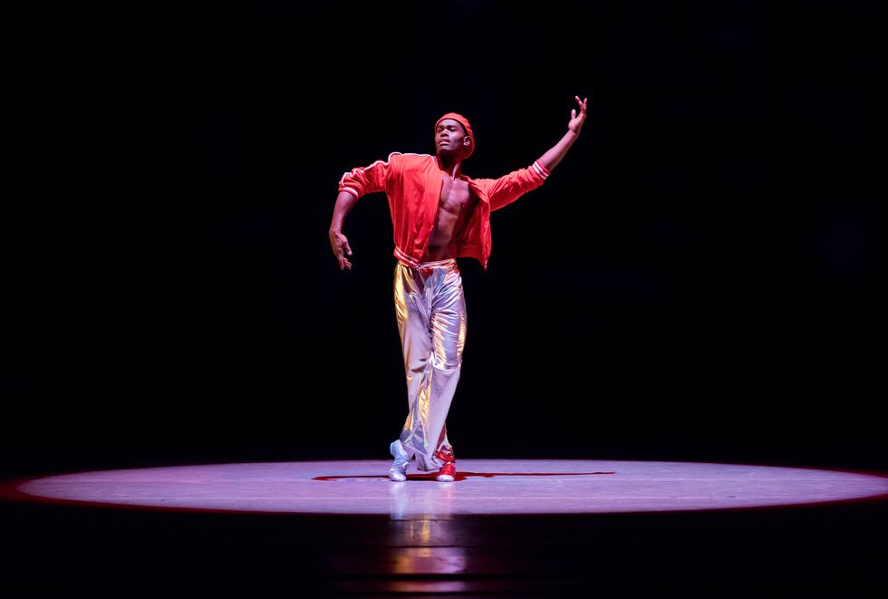Jamar Roberts onstage dancing in a spotlight. He wears silver pants, an open red jacket, exposing his bare chest, and a red hat. He has one foot crossed in front of the other, one arm extended upward in a semi circle shape, the other one extended downward.