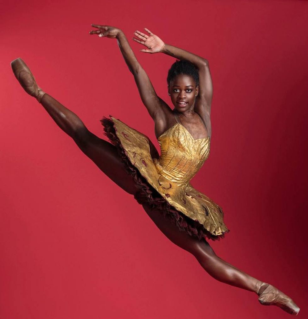 Michaela Deprince jumps against a red background at a photoshoot. She's wearing a gold costume with a classical tutu, her legs are straight and she looks directly at the camera, arms up and reaching behind her.