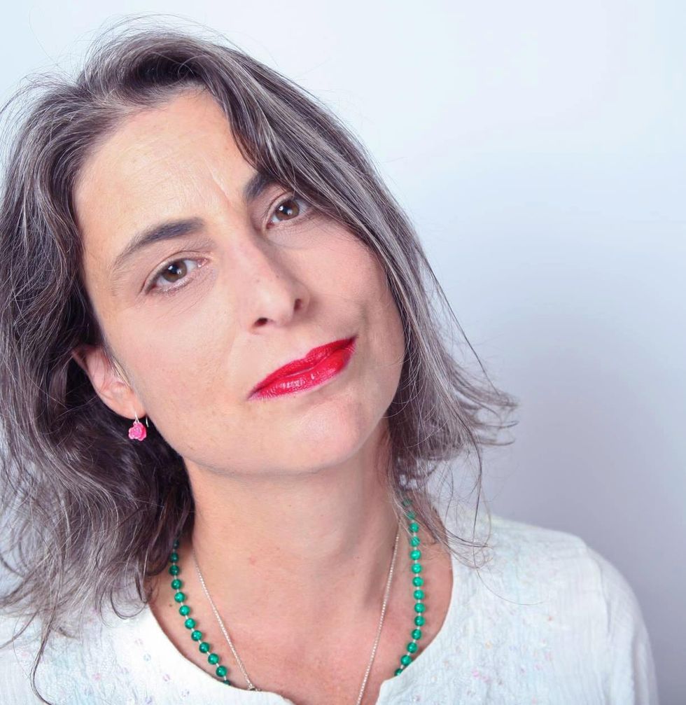 A grey haired woman with bright red lipstick and a green and silver necklace, pink drop earrings, tilts her hear to the side.