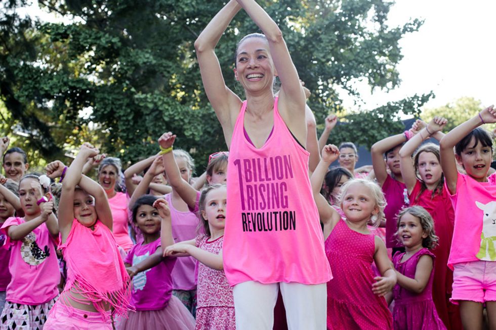 Charemaine seet claps her hands above her head, wearing a pink tank top that says "1 Billion Rising." she is surrounded by small girls in pink