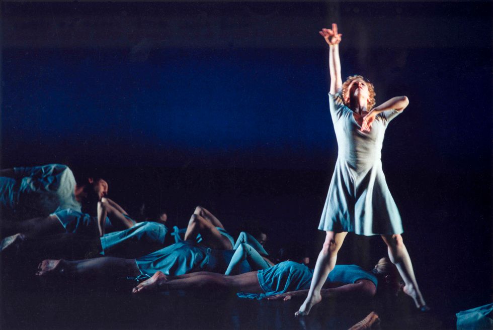 Lauren Grant stands in second position relevu00e9 downstage left in front of a row of dancers lying on the floor in a diagonal behind her.