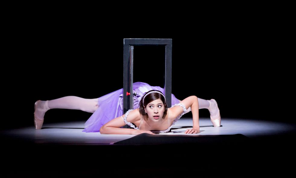 Beatriz Stix-Brunell in a bright purple dress lies on her stomach, legs stretched out to either side, with her torso squeezing through a miniature door frame.