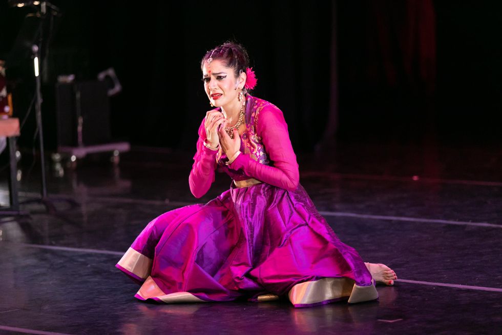 Rachna Nivas kneels on a stage in a bright pink dress, hands toward her heart and her expression strained